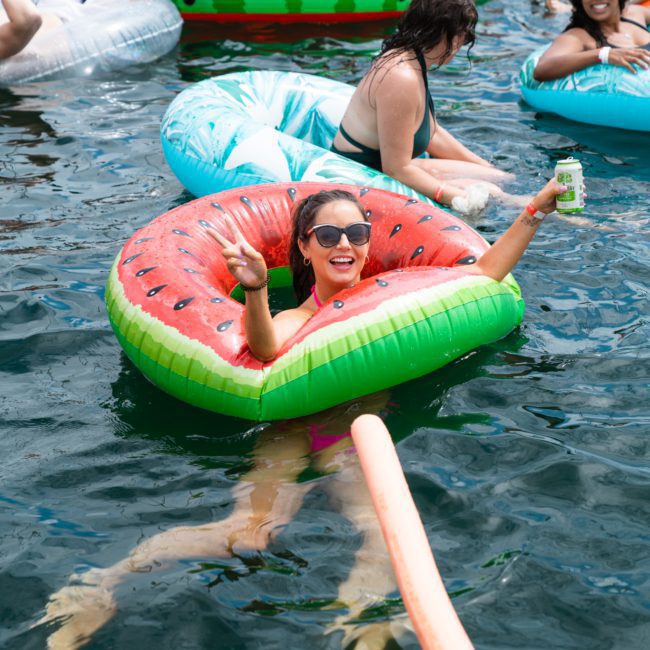 A group of individuals enjoying a day in the water on various inflatable floaties, with one woman prominently smiling and waving while holding a beverage can in a watermelon-shaped floatie during an exhilarating Sydney boat party hire.