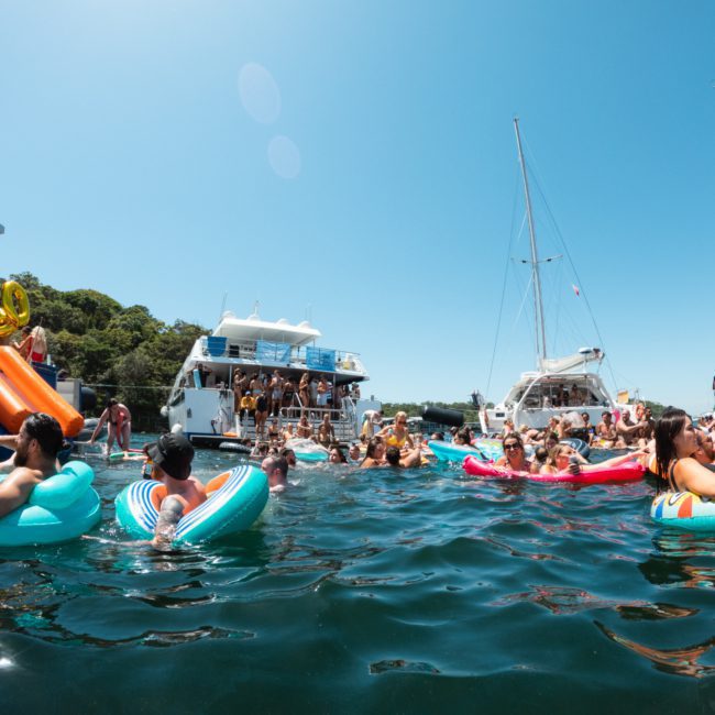 People relaxing on colorful inflatables in the water near several boats on a sunny day, enjoying a luxurious yacht hire in Sydney.