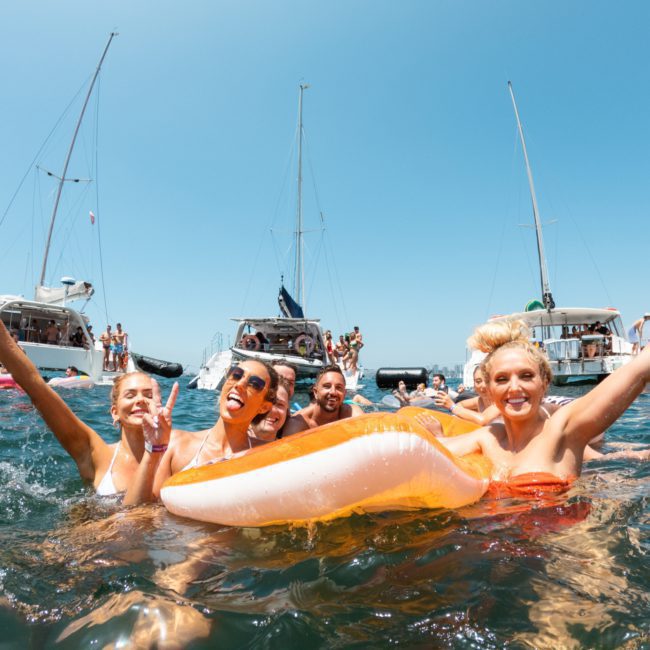 A group of people enjoying themselves in the water, holding onto inflatables, with several sailboats anchored in the background on a sunny day during a Sydney boat party hire.