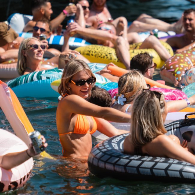 A group of people in swimsuits are enjoying a sunny day, lounging on inflatable tubes in a body of water, holding drinks and socializing during a luxurious catamaran party in Sydney.
