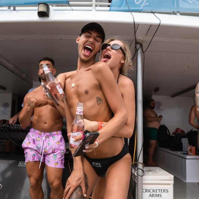 Two people in swimwear laugh and pose with bottled drinks on a boat. Other people and yacht club signs are visible in the background, adding to the festive atmosphere of a luxury yacht hire Sydney event.