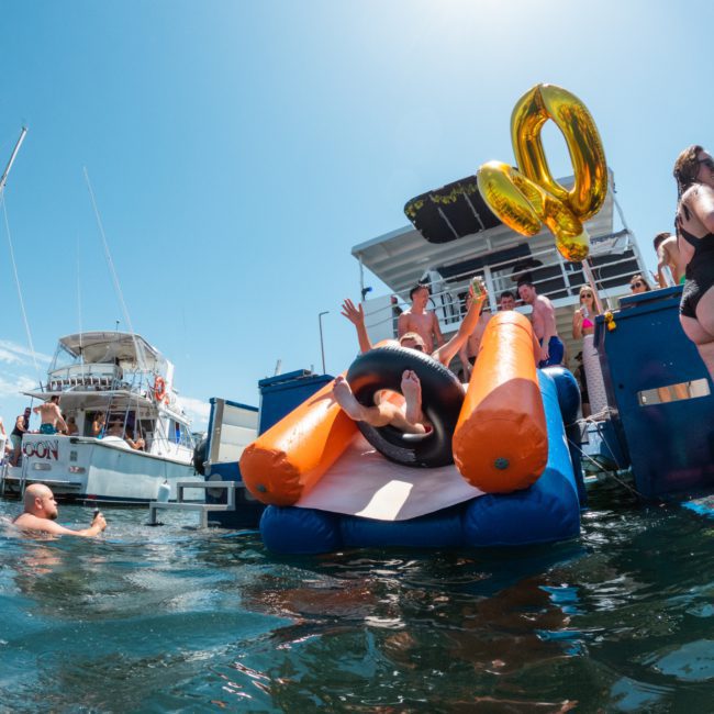People enjoying a sunny day on the water with inflated numbers "40," a person tubing down an inflatable slide, and boats in the background, capturing the essence of a luxury yacht hire Sydney experience.