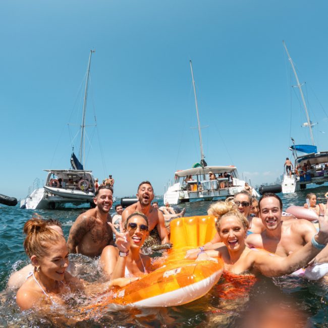 A group of people are enjoying a sunny day in the water surrounded by boats, smiling, and using floaties during a Sydney boat party hire.