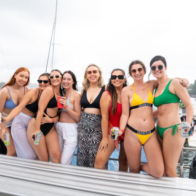 A group of eight women in swimwear stand on a boat, some holding drinks, with a body of water and trees visible in the background. Perfect for those considering a Luxury yacht hire Sydney for an unforgettable experience.