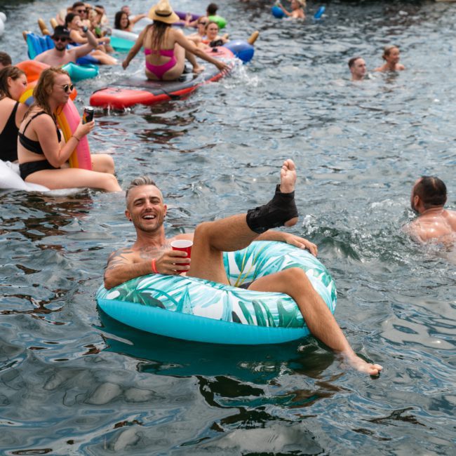 People are enjoying a sunny day in a body of water, floating on inflatable rings and swimming. One is holding a red cup and has a cast on his ankle. Others are socializing and relaxing in the background, perfect for imagining the ultimate catamaran party Sydney can offer.