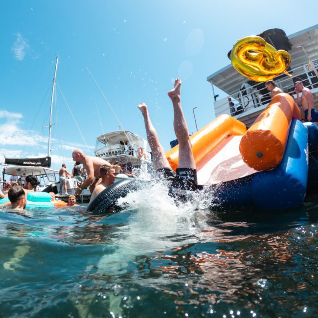 A person in shorts is mid-air after sliding off an inflatable slide into a body of water beside a large boat. Several people are swimming, and there is a large gold-colored inflatable number six floating nearby. Ideal for corporate boat events Sydney or luxury yacht hire Sydney, it's pure fun on the harbor.