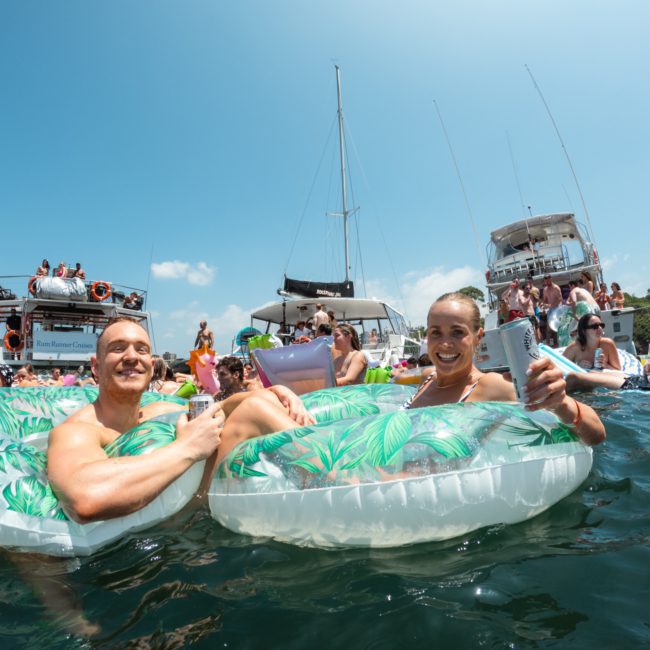 Two people float on inflatable rings with drinks in hand, surrounded by others in the water near boats under a clear sky, enjoying a catamaran party Sydney style.