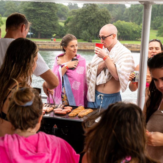A group of people, some wrapped in towels, gather around a barbecue grill with food cooking on it at an outdoor event near the water and greenery. Perfect for a catamaran party in Sydney, the atmosphere is filled with laughter and relaxation.