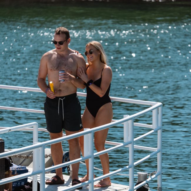 Group of people in swimwear standing and sitting on a white dock by the water on a sunny day. One man holds a drink while a woman beside him wears sunglasses and holds a beverage. Perfect setting for enjoying a luxury yacht hire Sydney or hosting an unforgettable catamaran party Sydney.