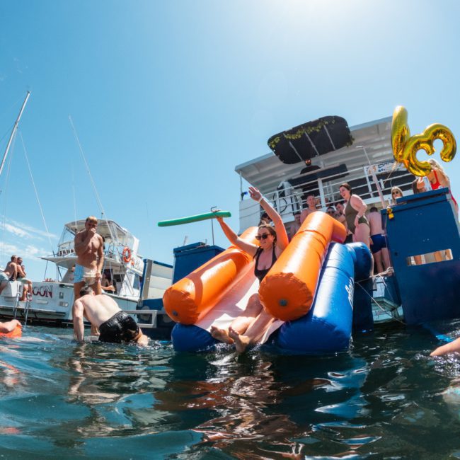 People enjoying a sunny day on the water, participating in various activities on an inflatable floating platform and nearby boats. The scene is lively with DJ boat hire Sydney adding vibrant music to the atmosphere, making it perfect for a memorable day.