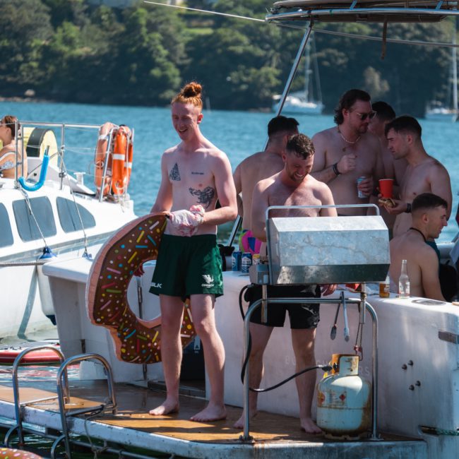 A group of people are on a boat deck near a grill, enjoying their time shirtless and one with a donut-shaped pool float. Other boats and a forested shoreline are visible in the background, setting the perfect scene for a lively Sydney boat party hire.