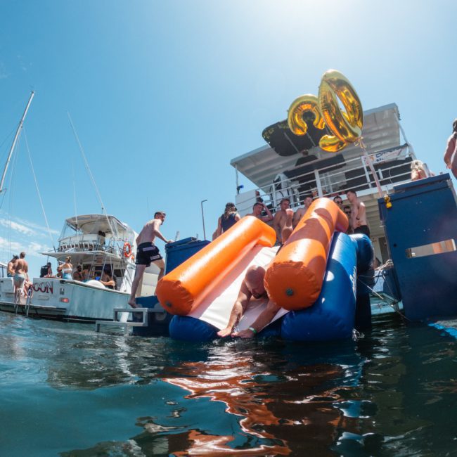 People enjoying a sunny day on a boat with orange inflatable floats and large gold "30" balloons in the background, with an adjacent luxury yacht hire Sydney nearby.