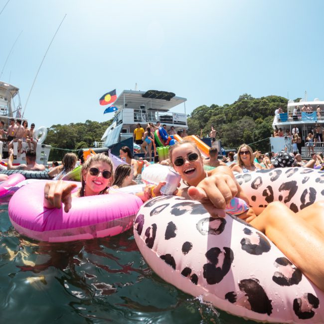 People are floating on inflatable tubes in the water near boats, enjoying a sunny day. Some are waving and pointing at the camera, with a luxury yacht hire Sydney adding an extra touch of elegance to the scene.