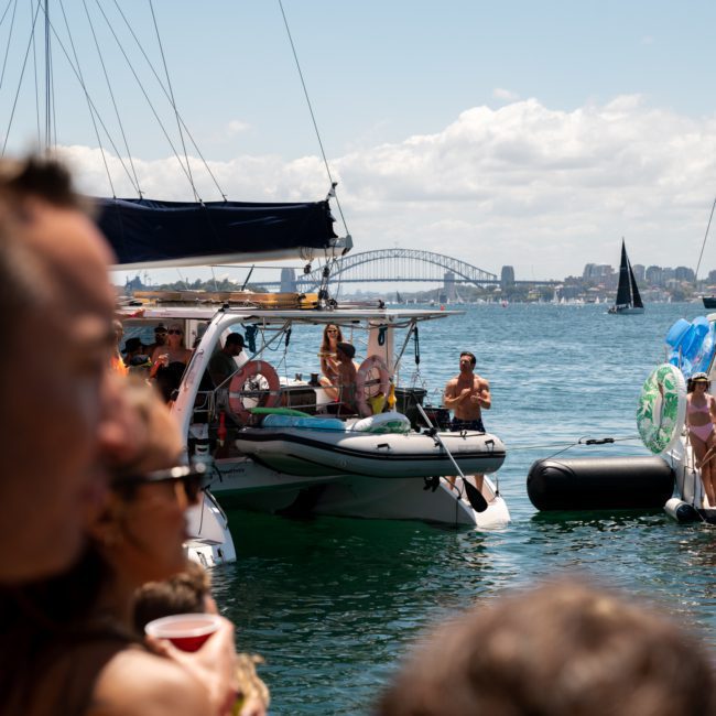 People enjoying a sunny day on boats near a harbor with a bridge in the background. Some are standing on decks while others float nearby. It’s an ideal scene for a private yacht charter or corporate boat events in Sydney Harbour.
