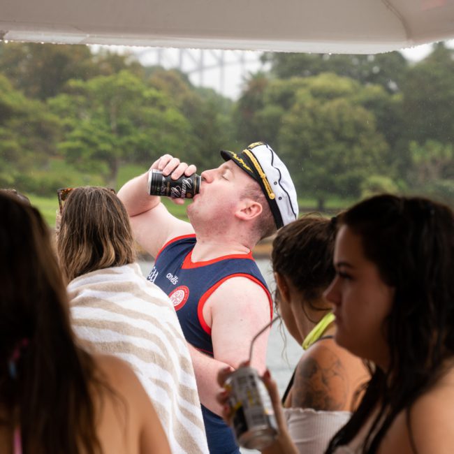 A group of people on a boat with one person wearing a captain's hat drinking from a can, surrounded by trees and water in the background, perfect example of Corporate boat events Sydney.