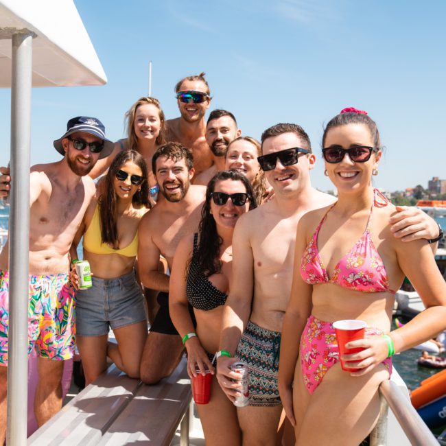 A group of people in swimsuits are posing together for a photo on a private yacht charter in Sydney Harbour. Some are holding drinks, and there are other boats in the water behind them. It's a sunny day.