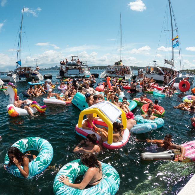 A crowded scene of people on various inflatable pool floats in the water, surrounded by boats on a sunny day, adds a lively touch to any Sydney boat party hire.