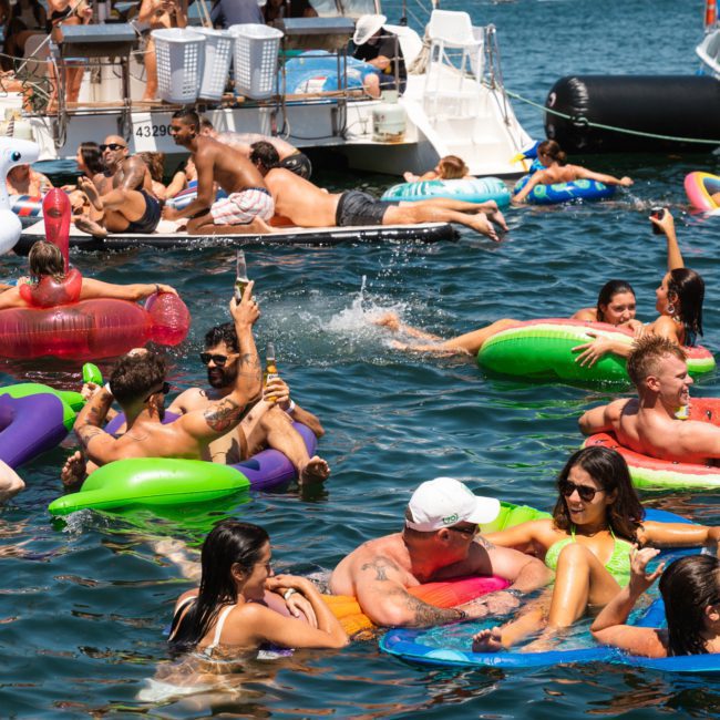 A group of people floating on various inflatables in the water near several boats on a sunny day, enjoying a lively Sydney boat party hire.