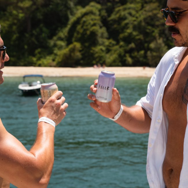Two people clink canned drinks on a catamaran party in Sydney, with a body of water and distant shoreline featuring greenery in the background.