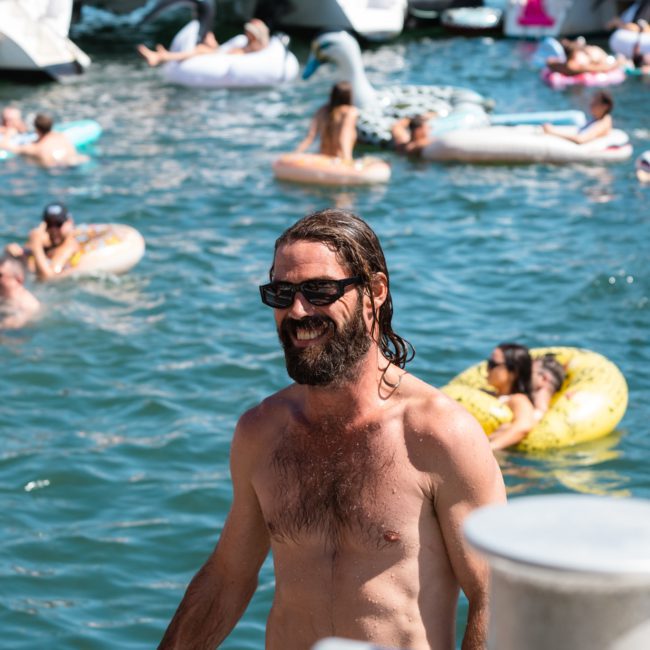 A bearded man with long hair and sunglasses is seen smiling, emerging from the water onto a boat. People are lounging on floaties in the water around various boats in the background, enjoying what appears to be a lively Sydney boat party hire.