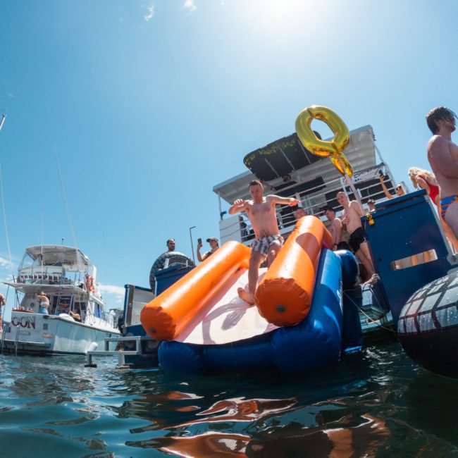 People on a boat enjoying a sunny day; a person is sliding down an inflatable slide into the water, creating perfect fun for a Sydney boat party hire.