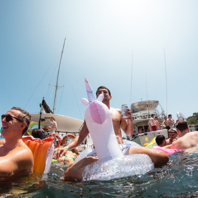 A group of people is enjoying a sunny day on the water, sitting on inflatables. A man on a unicorn float holds a drink. Boats are in the background, hinting at the luxurious vibe of a private yacht charter on Sydney Harbour.