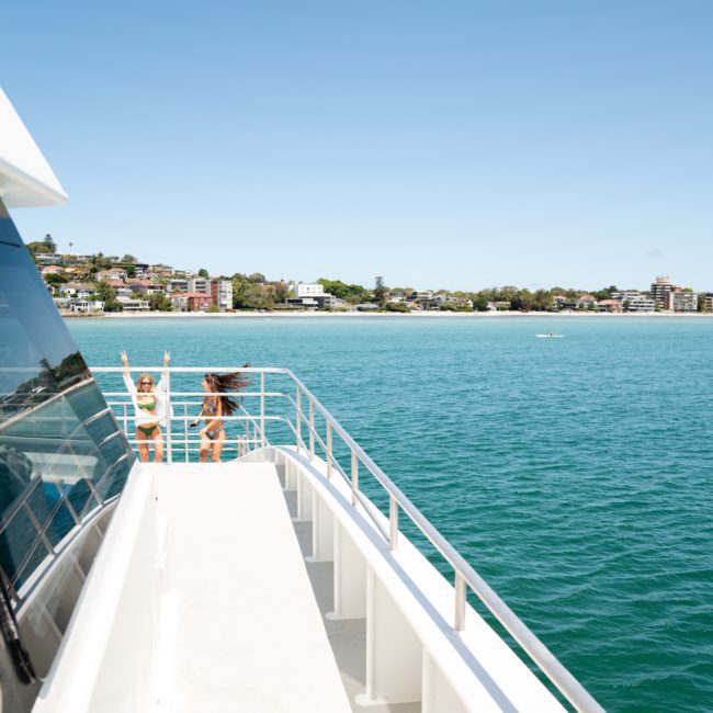Two individuals in swimsuits stand on the deck of a boat near the railing, with a coastal town and blue sea in the background, enjoying what seems like an exclusive luxury yacht hire in Sydney.