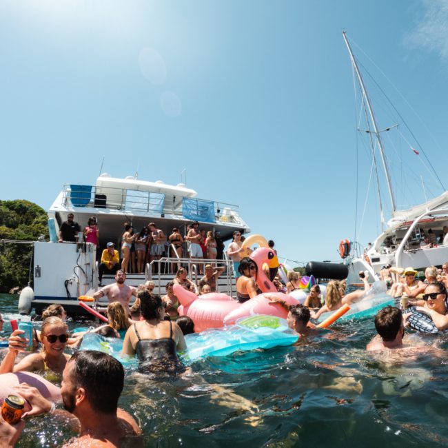 People are socializing and swimming with inflatable floats in the water near two anchored boats during a sunny day, enjoying a private yacht charter on Sydney Harbour.