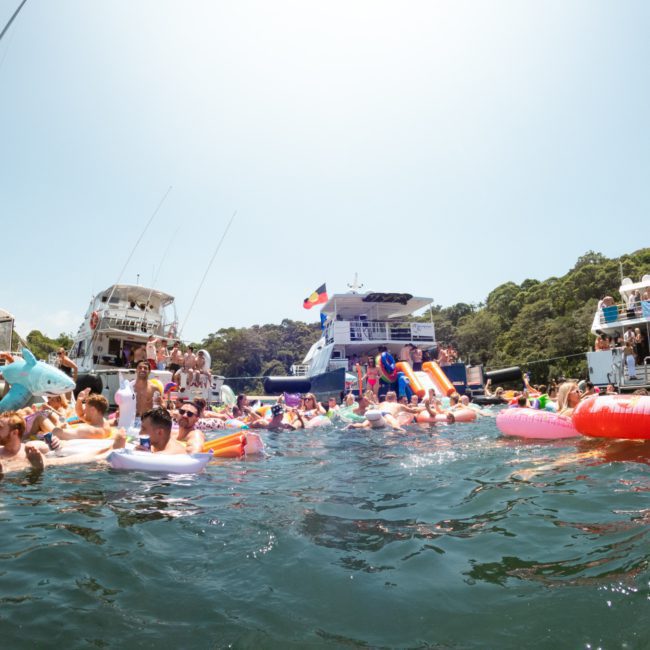 A large group of people are swimming and lounging on inflatables in a body of water, surrounded by several anchored boats on a sunny day. It looks like the perfect setting for a Sydney boat party hire or even corporate boat events Sydney.