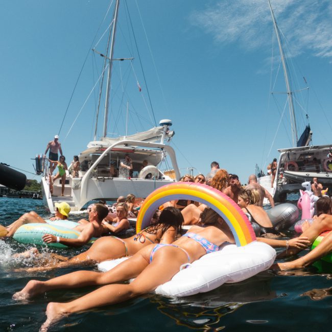 People on various inflatables enjoy a sunny day in the water next to anchored boats. The scene includes a rainbow float among other flotation devices, making it perfect for a Sydney boat party hire or corporate boat events Sydney.