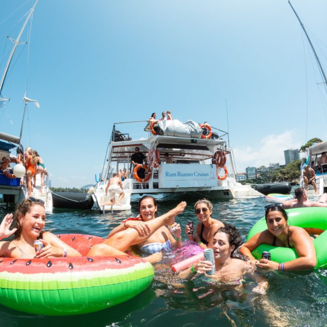 A group of people enjoying a sunny day on the water, floating on inflatable rings near a boat named "Rum Runner Cruises," with other boats and the distant shoreline visible. The scene feels like a catamaran party in Sydney, offering a fun and vibrant experience for everyone.