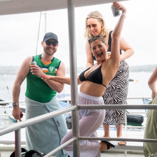 Three people on a boat, one man with a drink smiling, and two women: one energetically posing in front and the other holding a can behind them. Perfect scene for DJ boat hire Sydney or an unforgettable Catamaran party Sydney.
