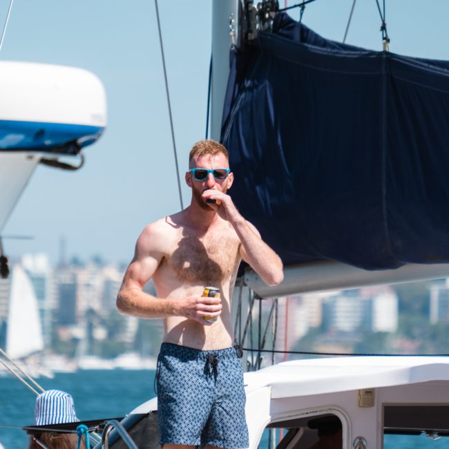 A person in sunglasses and swim shorts stands on a sailboat, holding a beverage, with buildings visible in the background, enjoying a luxury yacht hire Sydney experience.
