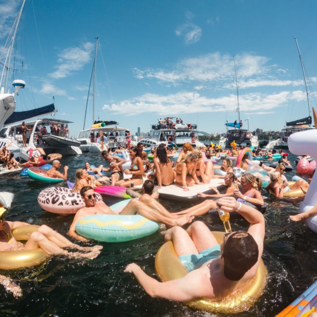 A vibrant scene of people on various inflatable floats in the water between docked boats, enjoying a sunny day with clear skies during a luxury yacht hire in Sydney Harbour.