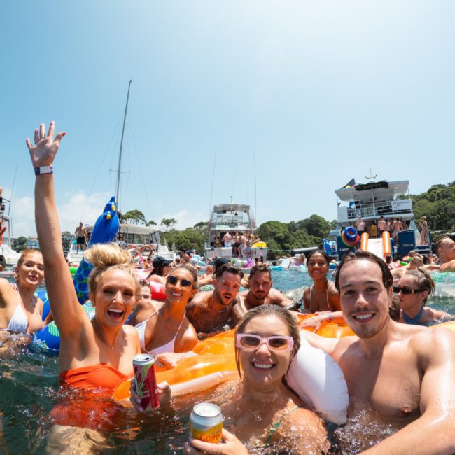 A group of people in swimsuits are enjoying a sunny day in the water at a catamaran party Sydney. They're surrounded by boats and floatation devices. Some are holding drinks, and one person is waving towards the camera.