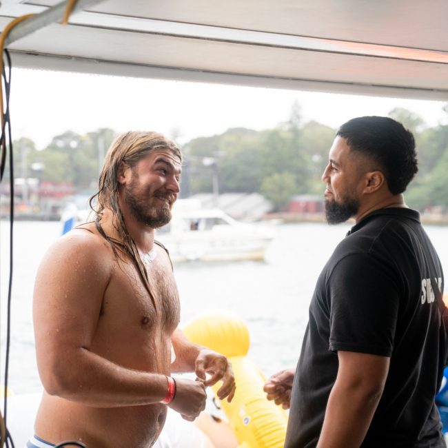 Two men are conversing on a boat; one is shirtless and wet, while the other wears a black shirt labeled "SECURITY." Inflatable toys and another boat are in the background, suggesting an exciting corporate boat event in Sydney.