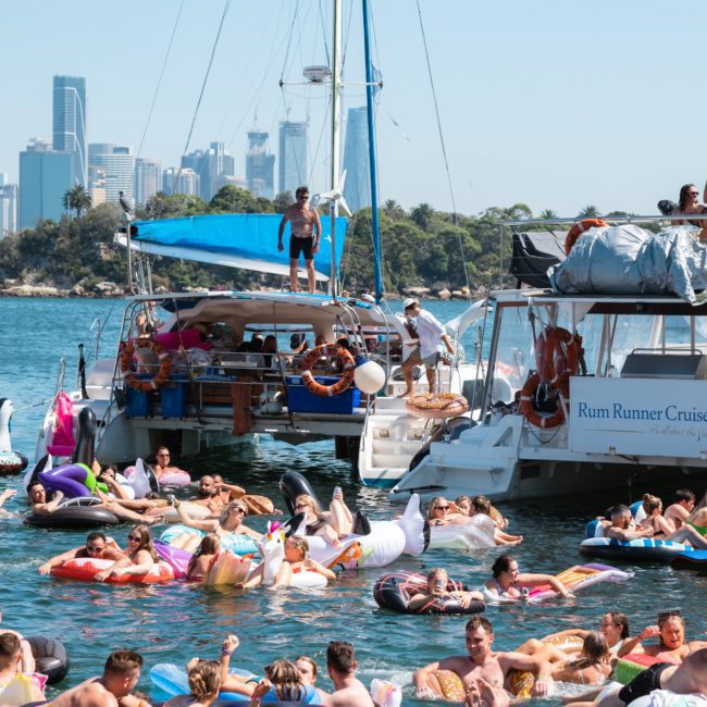 People relax on inflatable floats in the water next to anchored yachts on a sunny day, with a city skyline visible in the background, enjoying the benefits of a private yacht charter Sydney Harbour.