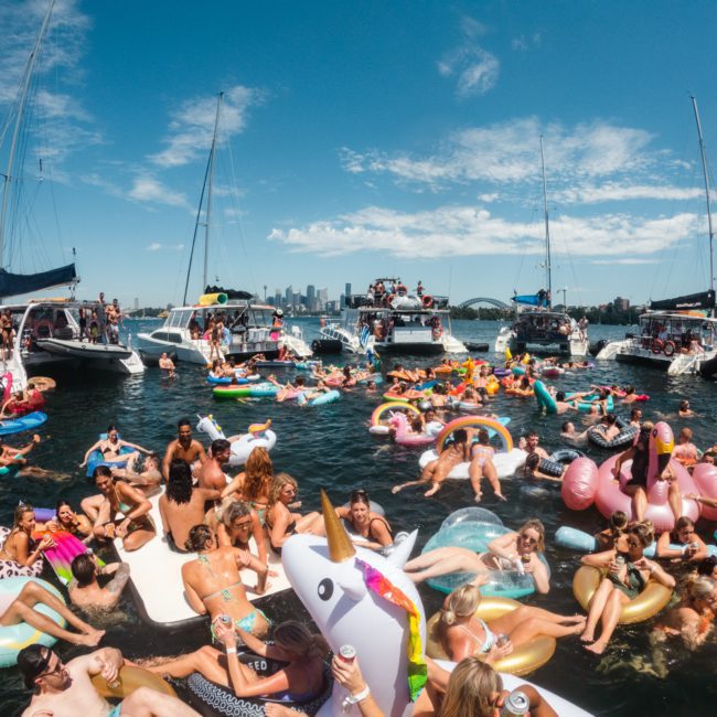 A large group of people on inflatable floats and boats enjoying a sunny day on the water with the city skyline visible in the background, highlighting a luxury yacht hire Sydney experience.