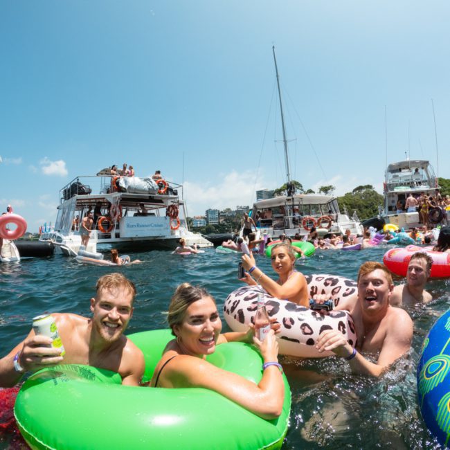 People enjoying a sunny day on the water, swimming and lounging on inflatable floats near several anchored boats at a Sydney boat party hire event.