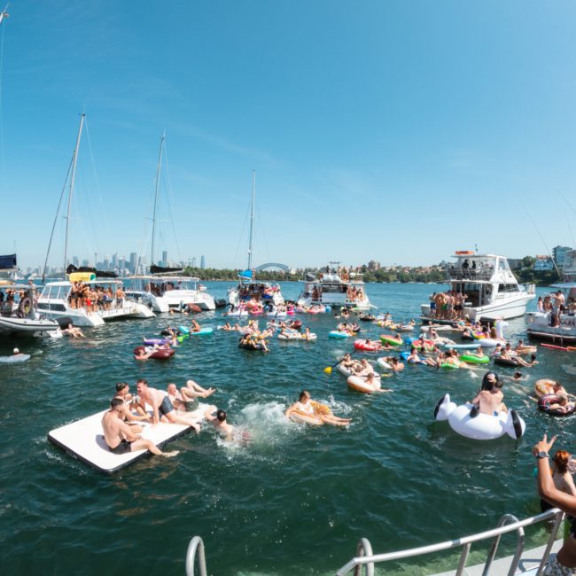 People are enjoying a sunny day on the water, swimming and floating on inflatables between anchored boats during a catamaran party in Sydney.