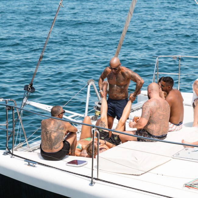 A group of people with visible tattoos relaxes and socializes on the deck of a white sailboat on a sunny day, enjoying a private yacht charter in Sydney Harbour.
