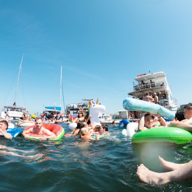People are floating in the water on inflatable rafts and floats near several boats on a sunny day, enjoying a Catamaran party Sydney experience.