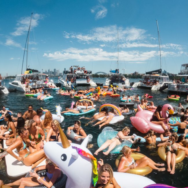 A large group of people socialize on various inflatable pool floats, including a unicorn, in a body of water surrounded by boats on a sunny day. Consider enhancing your experience with a private yacht charter Sydney Harbour for an unforgettable time.