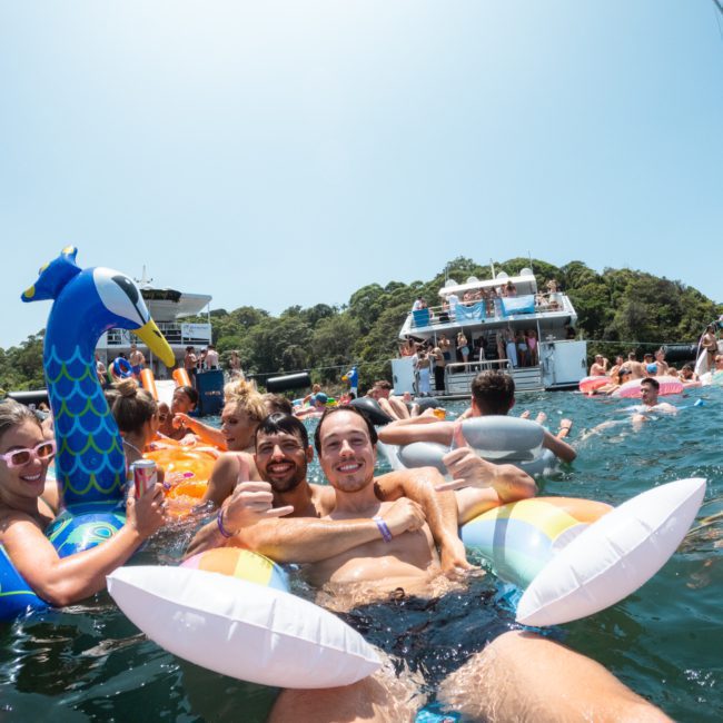 People enjoying a party on the water, some in colorful inflatable rafts, with boats and trees in the background. Experience luxury yacht hire Sydney or catamaran party Sydney for an unforgettable time.