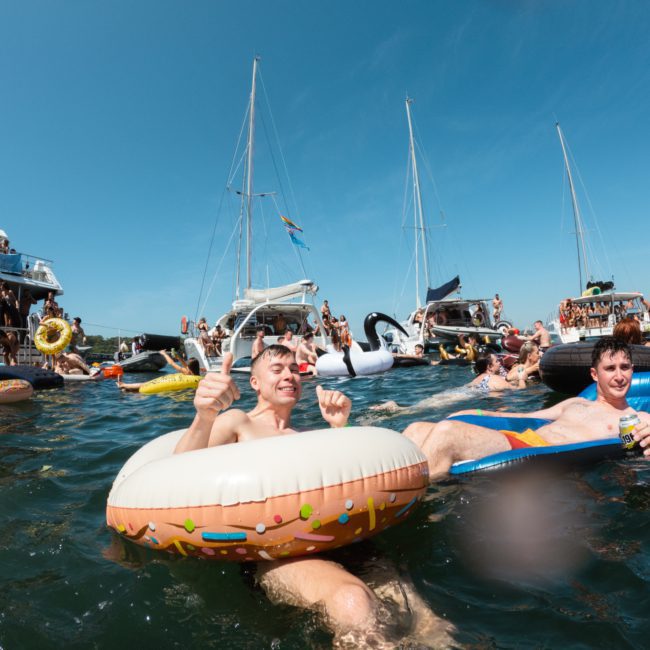 Two people float on inflatables in a crowded, festive water scene with multiple boats in the background under a clear blue sky, highlighted by a private yacht charter on Sydney Harbour.
