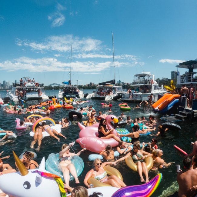 A crowd of people float on colorful inflatables in a busy waterway, surrounded by boats under a bright blue sky, enjoying the vibrant scene reminiscent of a Sydney boat party hire.