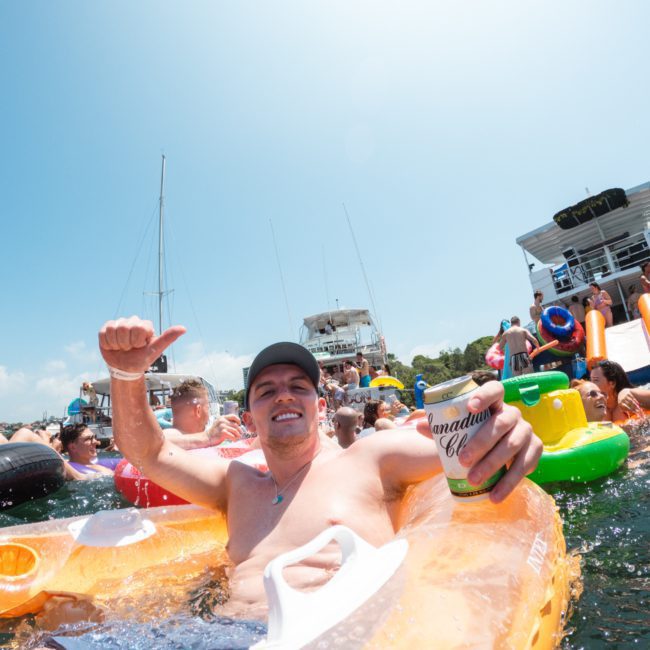 A man is floating in a lake on an inflatable raft holding a beverage, surrounded by other individuals on inflatables and boats in the background, evoking the vibe of a Sydney boat party hire.