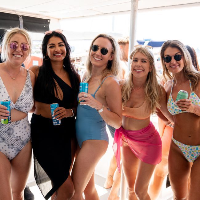 Five women in swimsuits standing together on a boat, smiling and holding canned drinks, during a catamaran party in Sydney. Other people and boats are visible in the background, creating a lively atmosphere.