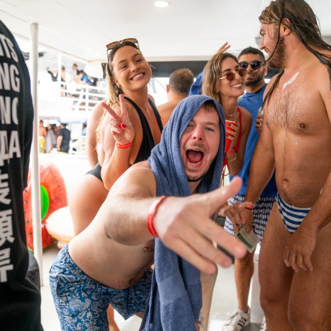 A group of people in swimwear are gathered at a party. One man in the foreground, wrapped in a towel, gestures enthusiastically towards the camera while others smile and pose behind him, enjoying their Catamaran party Sydney adventure.