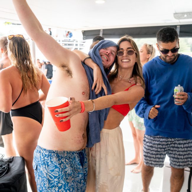 Group of people on a private yacht charter on Sydney Harbour, smiling and celebrating. A man with a blue towel on his head and a woman hugging hold red cups. Others around them are casually dressed, holding drinks, enjoying the water in the background.